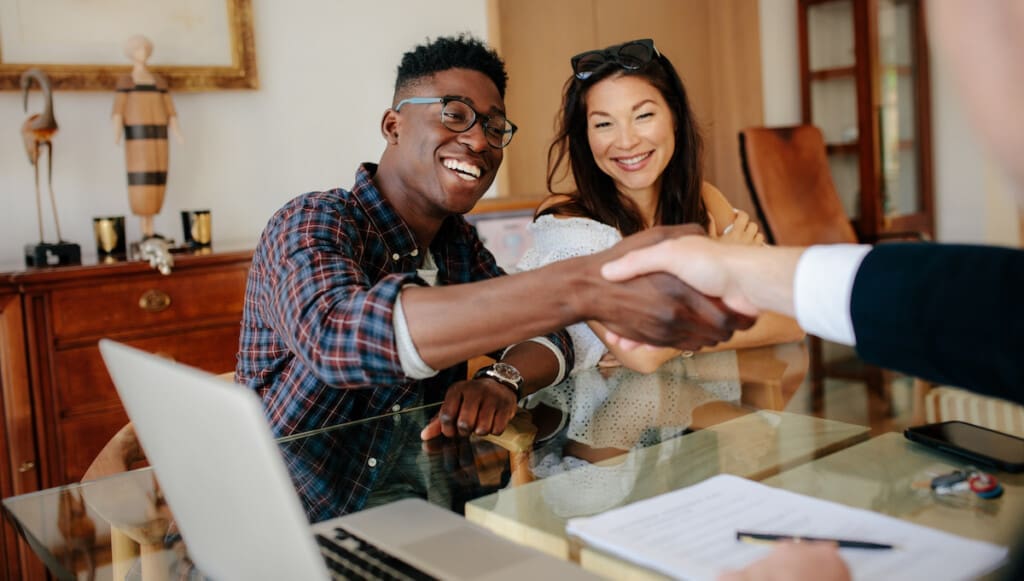 A smiling couple sits at a glass table, with the man shaking hands with another person across from them finalizing the purchase of their new Holt Home.