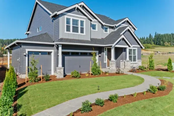 A two-story gray house with white trim and a black roof sits gracefully near the Green Mountain glades. It features a double garage, large windows, and a neatly landscaped yard with a curved concrete path and young trees under the clear blue sky.