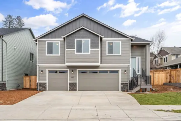 A modern two-story gray house with white trim, three-car garage, and a small porch with stairs. The driveway is concrete, and the yard is bordered by mulch and grass under a partly cloudy sky.