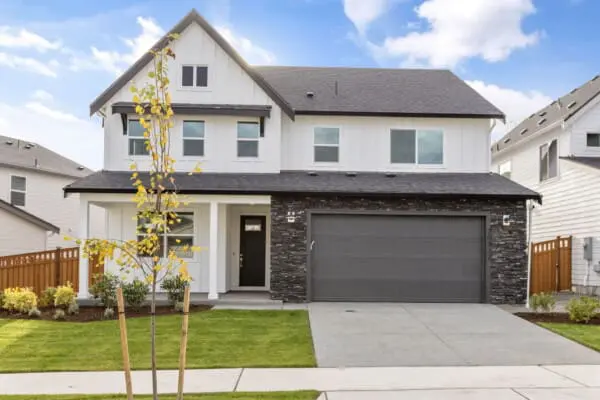 Modern two-story house with white siding and dark stone accents, a dark gray garage door, a small front yard with green grass, young trees, and shrubs, set under a blue sky with scattered clouds.