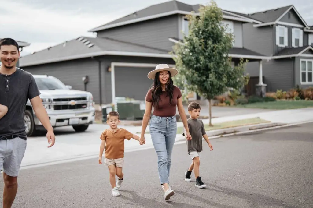 A woman holding hands with two young boys walks across a suburban street, while an adult man walks nearby. The group is in front of modern gray houses on a cloudy day.