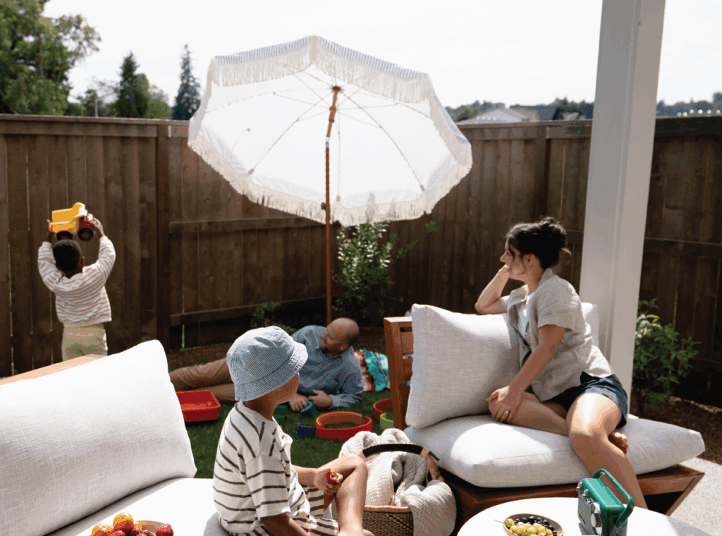 A family enjoys a sunny day in a backyard. A child plays with a toy truck near a wooden fence, while an adult and another child relax on a blanket. A woman lounges on an outdoor sofa under a large white umbrella.