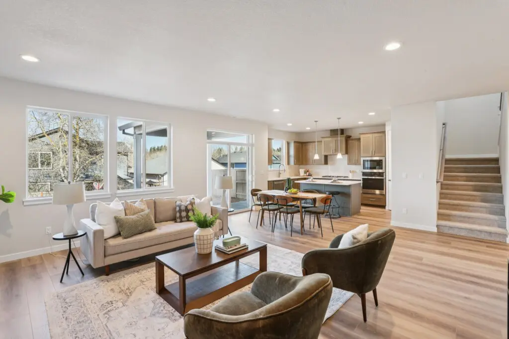 Open-concept living room, dining area, and kitchen with light wood floors, beige walls, modern furniture, and a large rug at 17269 SW Atfalati Lane; natural light pours in from several windows with the staircase visible on the right.