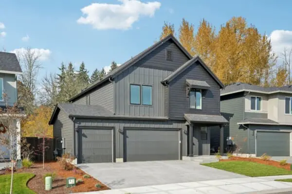 A modern two-story dark gray house by Alder & Ash at 2242 Ash St features a three-car garage, concrete driveway, and manicured lawn, surrounded by trees with autumn foliage under a blue sky with scattered clouds.