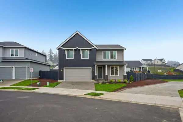 A modern two-story gray house with white trim and a double garage, set on a clean suburban street with manicured lawns and neighboring homes in the background under a clear blue sky.