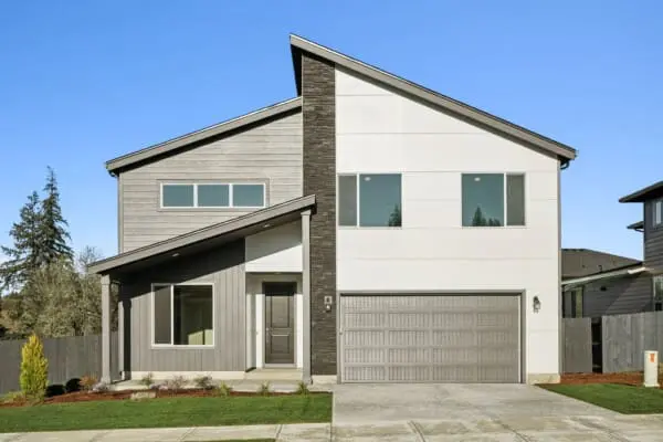 Modern two-story house with contrasting gray and white siding, large windows, a slanted roof, and a double garage. The front yard features grass, a small bush, and a concrete driveway under a clear blue sky.