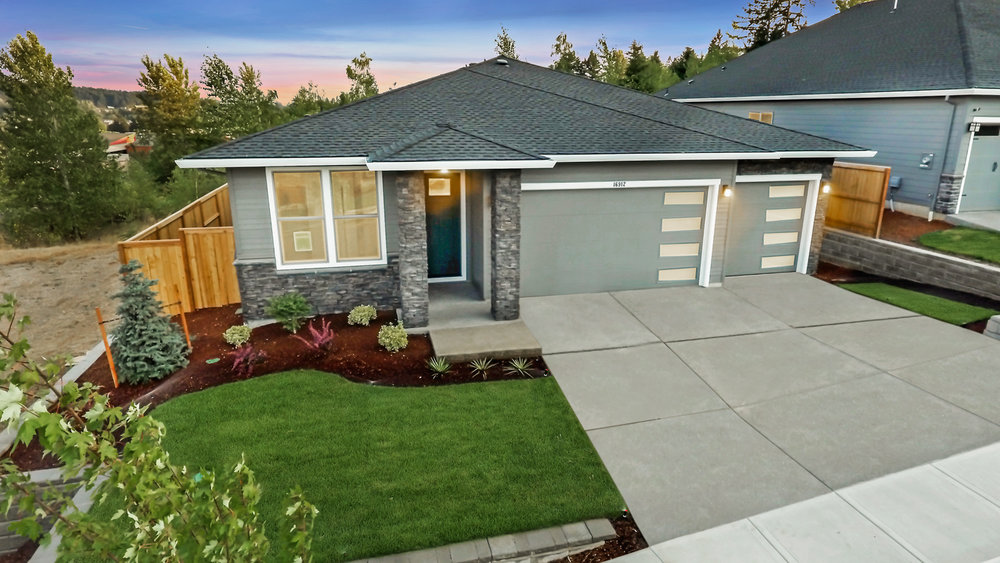A modern single-story house with a gray facade, two-car garage, and stone accents. It features a neatly landscaped front yard with green grass, shrubbery, and a small tree. The background shows trees and a colorful sunset sky.