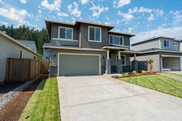 A modern two-story house with gray siding, large windows, and a double garage sits on Scouters 325 SE Webelos Way. The yard features neatly trimmed grass and landscaping, flanked by similar homes under a partly cloudy sky.