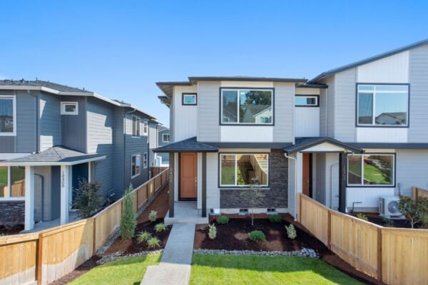 A modern two-story townhouse at 18406 NE 43rd Place in Ramble Creek features gray siding, large windows, a red front door, and a small landscaped front yard enclosed by a wooden fence on a sunny day.